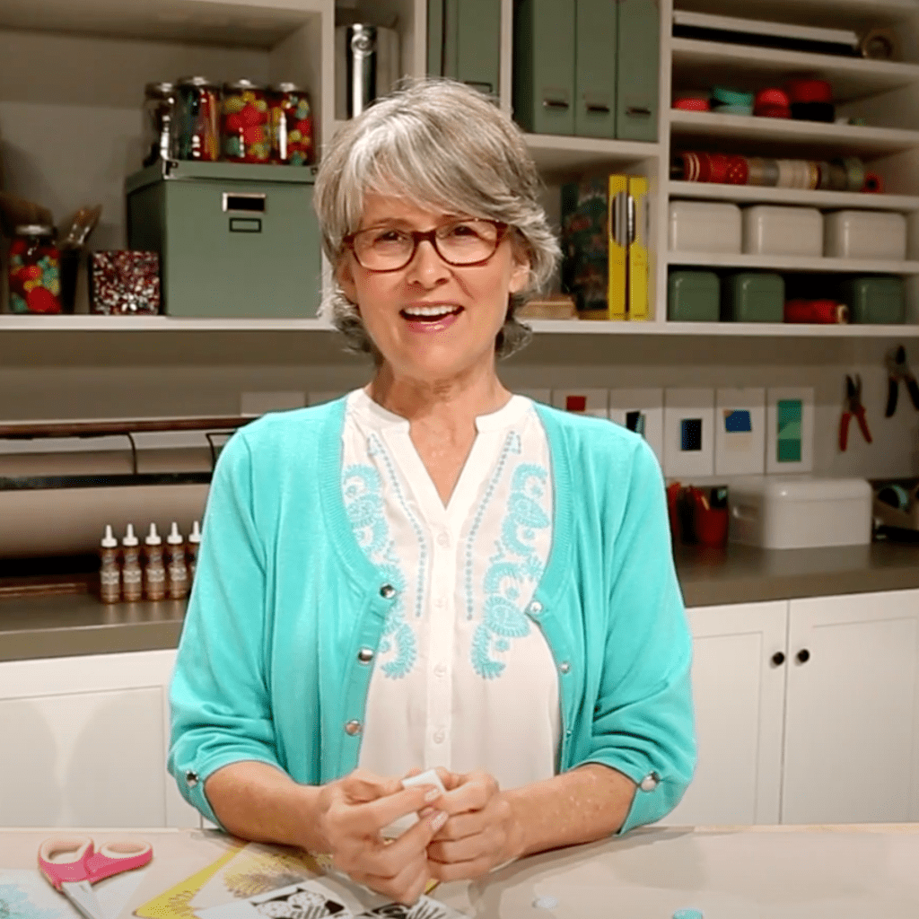 A smiling woman with gray hair is wearing a turquoise cardigan and holding crafting materials on a table in a well-organized creative workspace.
